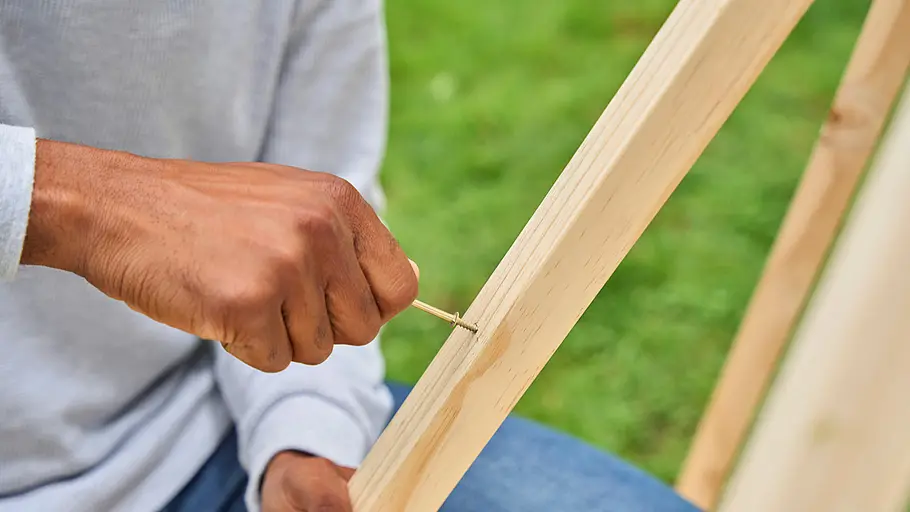 Eine Person dreht von Hand eine Schraube in eine aufrecht stehende Holzlatte des Tipi-Gestells auf einer Wiese.