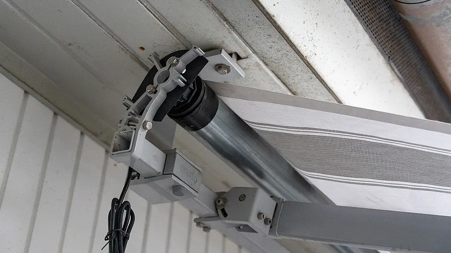 Montierte Markisenwelle Eine Markisenwelle mit montiertem Motor ist an einer Metallhalterung unter einem Dachüberstand befestigt.