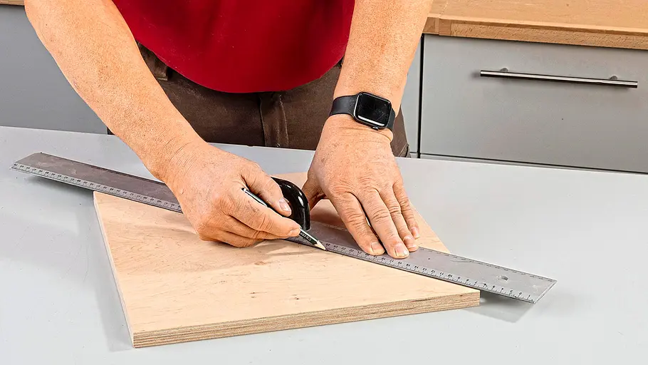 Mittelpunkt markieren Eine Person zeichnet mit einem Bleistift und Metallwinkel Linien auf eine quadratische Holzplatte.