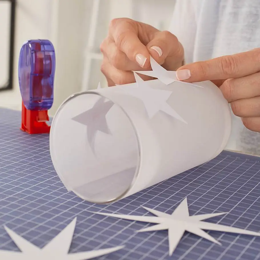 Weihnachts-Windlicht: Funkelndes Sternen-Glas Sterne aus Papier werden von einer Person auf ein mit Transparentpapier umwickeltes Glas geklebt.