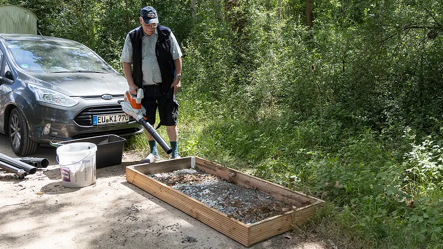 Ein Mann steht neben einem Auto und testet mit einem Laubsauger eine mit Laub und Steinen gefüllte Holzkiste am Waldrand.