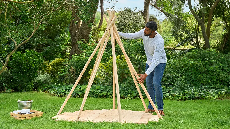 Ein Mann richtet auf einer Rasenfläche Holzlatten zu einem Tipi-Gestell auf einer Holzplattform aus.