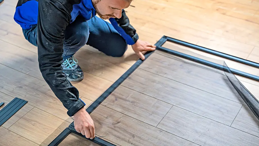 Eine Person kniet auf einem Holzfußboden und befestigt ein Schutzgitter im schwarzen Rahmen einer Insektenschutztür.