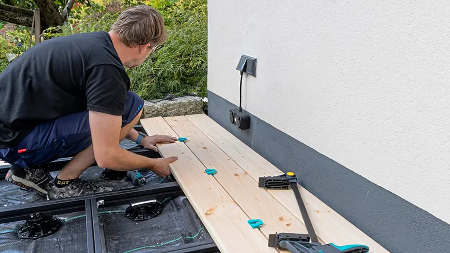 Terrasse gestalten Ein Mann legt Holzdielen auf eine Terrassenunterkonstruktion und richtet sie mit Abstandhaltern und Zwingen aus.