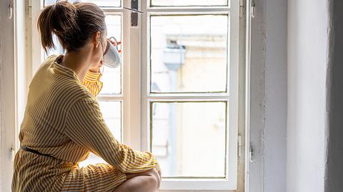 Frau sitzt am Fenster mit Tasse - Foto: iStock/StockRocket