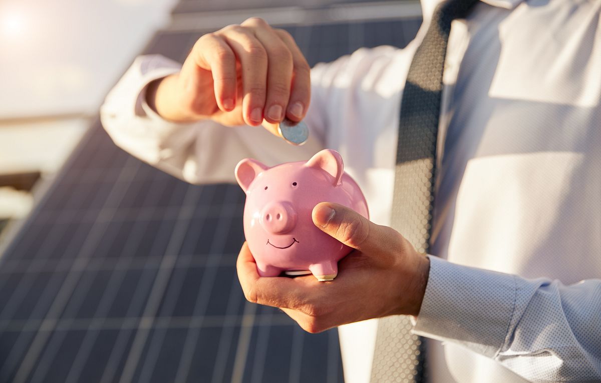 Ein Mensch steckt Kleingeld in ein rosa Sparschwein, im Hintergrund sind Solarpanels zu sehen