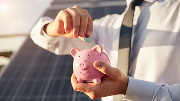 Ein Mensch steckt Kleingeld in ein rosa Sparschwein, im Hintergrund sind Solarpanels zu sehen - Foto: istockphoto.com / max-kegfire Ein Mensch steckt Kleingeld in ein rosa Sparschwein, im Hintergrund sind Solarpanels zu sehen - Foto: istockphoto.com / max-kegfire