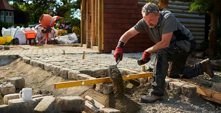 Ein Mann kniet im Garten neben einem neu angelegten Weg aus Natursteinen. Mit roten Arbeitshandschuhen verteilt er Sand mit einer Kelle und prüft mit einer Wasserwaage die Ausrichtung der Pflastersteine. Neben ihm liegen Werkzeuge, im Hintergrund stehen Baumaterialien und ein Betonmischer.