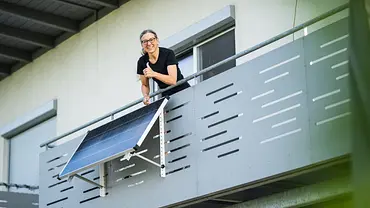 Eine Frau mittleren Alters steht lachend auf einem Balkon und zeigt Thumbs Up, am Balkon hängt ein Solarpanel - Foto: istockphoto.com / amriphoto Eine Frau mittleren Alters steht lachend auf einem Balkon und zeigt Thumbs Up, am Balkon hängt ein Solarpanel - Foto: istockphoto.com / amriphoto