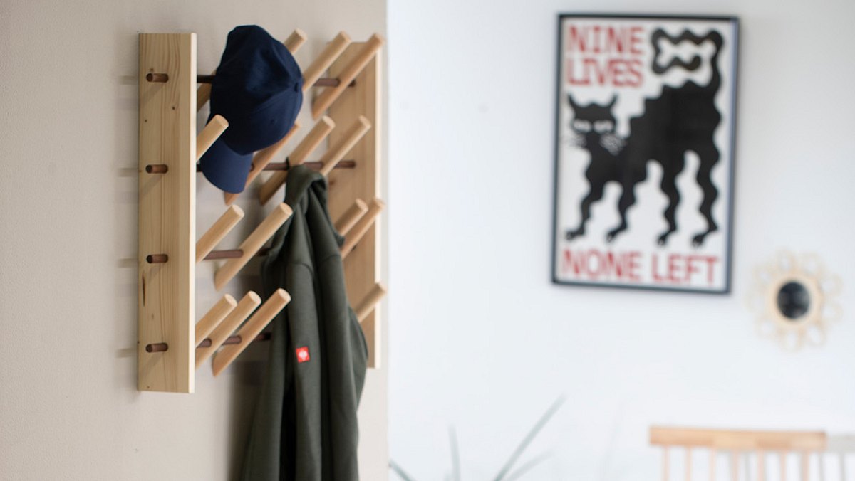 Eine Holz-Garderobe mit schräg stehenden Haken, an denen eine blaue Kappe und eine grüne Jacke hängen.