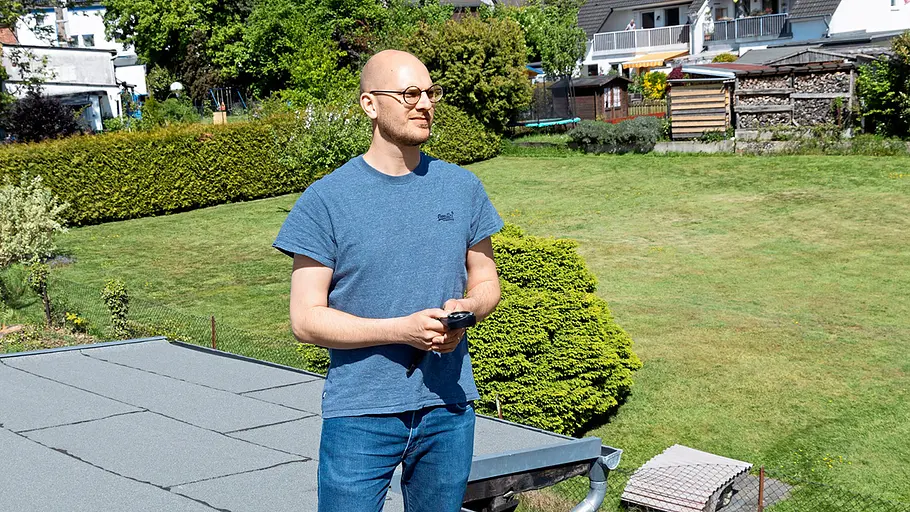 Kleine Solaranlage Ein Mann mit Brille steht auf einem Flachdach und blickt in einen sonnigen Garten, er hält einen Kompass.