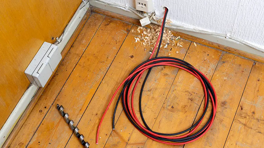 Kleine Solaranlage Zwei Kabelstränge, Bohrer und Holzspäne liegen am Holzboden, die Kabel verlaufen durch ein Wandloch ins Zimmer.