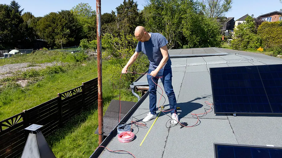 Kleine Solaranlage Ein Mann steht auf einem Flachdach und verlegt rote sowie schwarze Kabel, im Hintergrund Garten und Solarpanels.