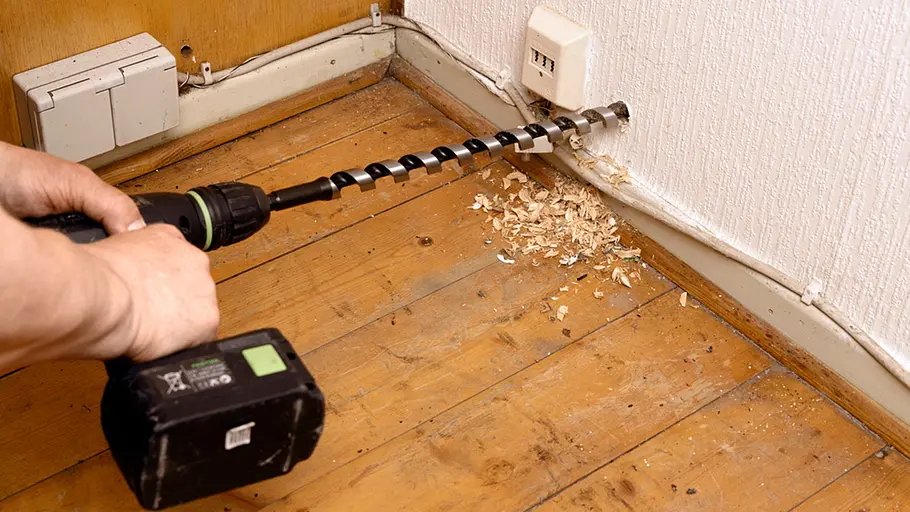 Kleine Solaranlage Mit einer Bohrmaschine wird ein Loch durch eine Zimmerwand nahe dem Boden gebohrt, Holzspäne liegen daneben.