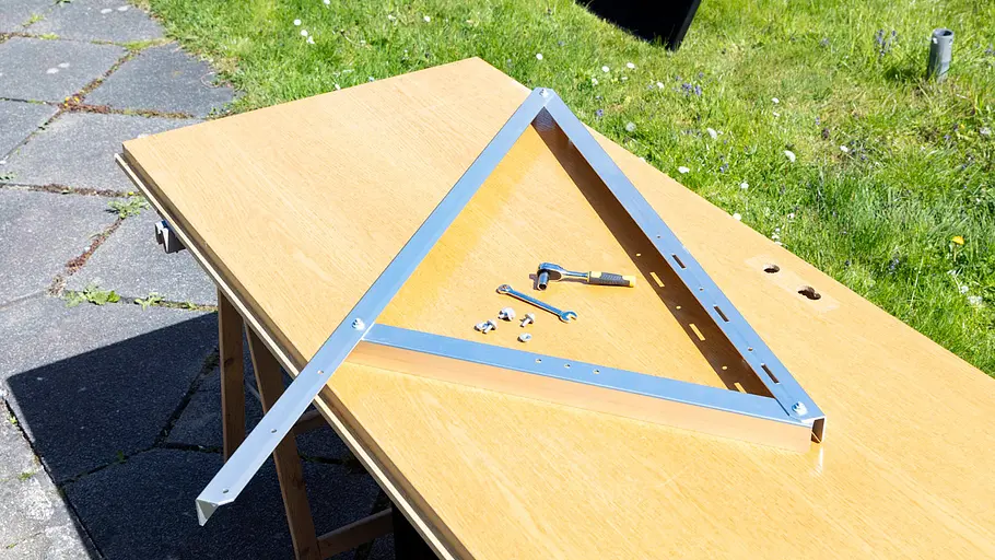 Kleine Solaranlage Ein Metallwinkelgestell, Schrauben und Werkzeuge liegen auf einem Holztisch im Freien neben einer Rasenfläche.