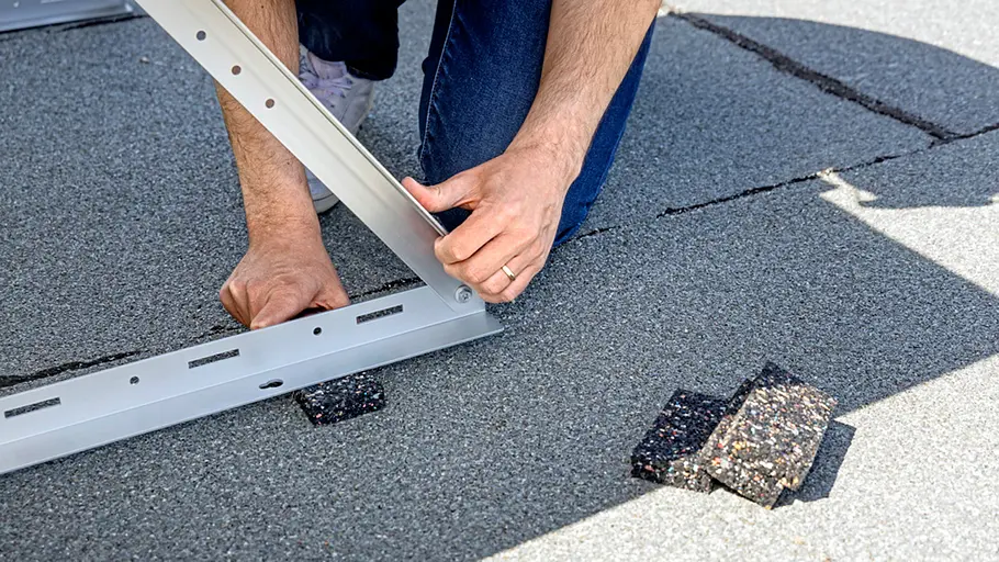 Kleine Solaranlage Eine Person richtet ein Metallgestell auf einem Flachdach aus, untergelegt mit schwarzen Gummiplatten-Abschnitten.