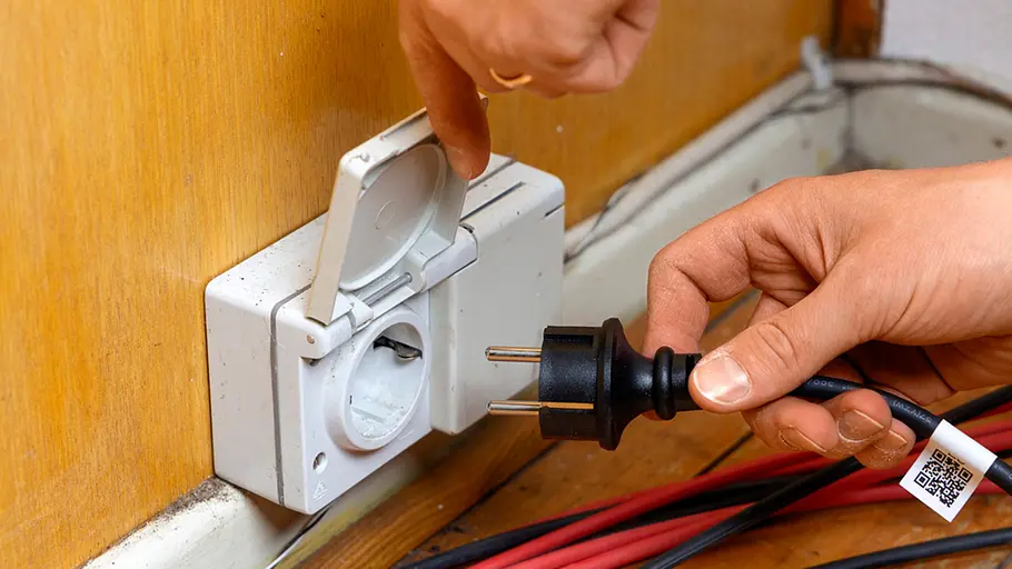 Kleine Solaranlage Eine Hand hält einen Stecker vor eine geöffnete Steckdose am Boden, rote und schwarze Kabel verlaufen daneben.