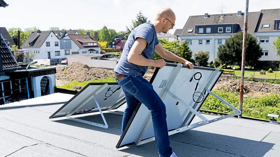 Kleine Solaranlage Ein Mann richtet auf einem Flachdach zwei Solarpanels mit Metallgestellen aus, im Hintergrund Häuser und Gärten.
