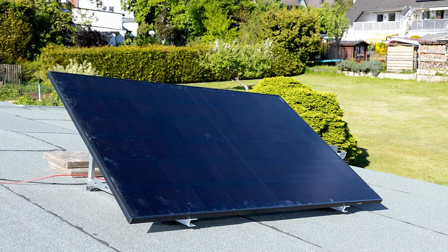 Kleine Solaranlage Ein Solarpanel steht schräg auf einem Flachdach im Sonnenlicht, im Hintergrund Garten und umliegende Häuser.
