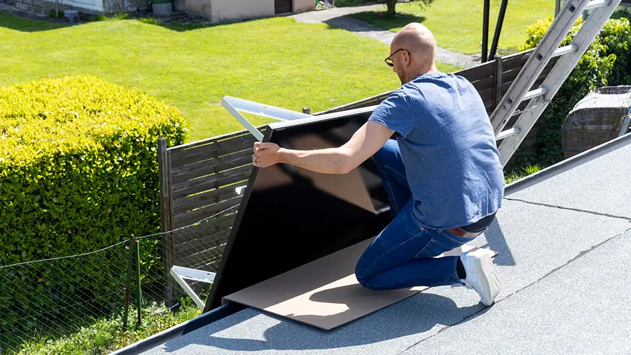 Kleine Solaranlage Ein Mann kniet auf einem Flachdach und richtet ein Solarpanel mit Halterung aus, im Hintergrund Garten und Leiter.