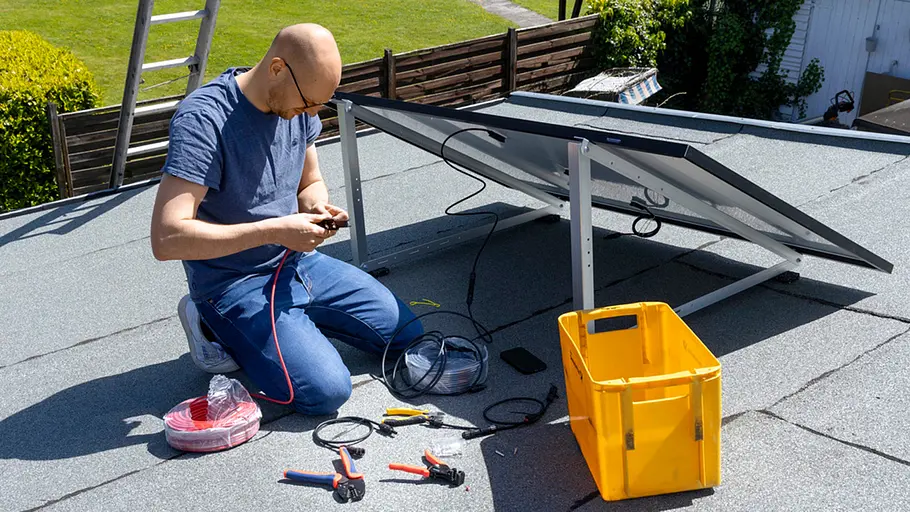 Kleine Solaranlage Ein Mann kniet auf einem Flachdach, verbindet ein Solarkabel mit Werkzeug neben einem schräg aufgestellten Solarpanel.