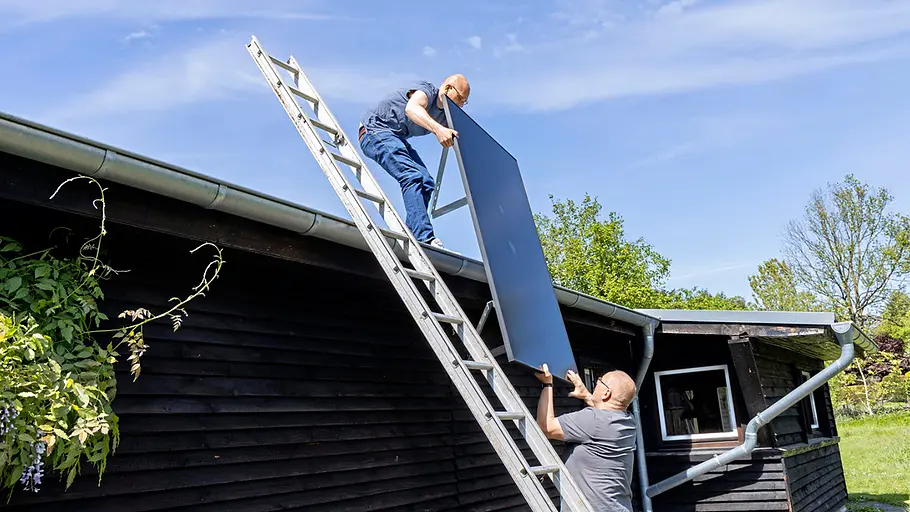 Kleine Solaranlage Zwei Männer heben gemeinsam ein Solarpanel über eine Leiter auf das Dach eines dunklen Gartenhauses bei sonnigem Wetter.