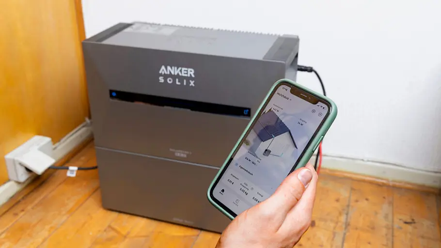 Kleine Solaranlage Eine Hand zeigt eine App auf dem Smartphone, im Hintergrund steht der Energiespeicher Anker Solix am Boden.