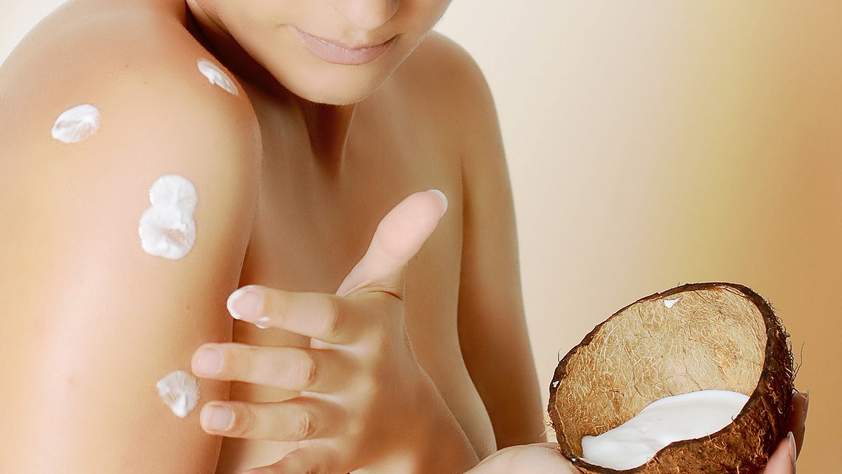 Kokosöl eincremen Eine Person trägt Kokosöl auf ihre Schulter auf und hält eine halbe Kokosnuss mit weißer Creme darin.
