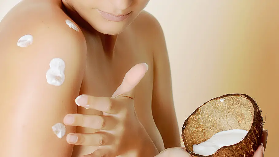 Kokosöl eincremen Eine Person trägt Kokosöl auf ihre Schulter auf und hält eine halbe Kokosnuss mit weißer Creme darin.