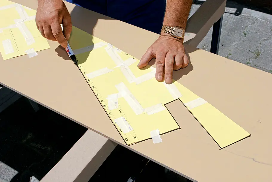 Eine Hand zeichnet eine Kontur auf Holz anhand einer angelegten Pappschablone nach.