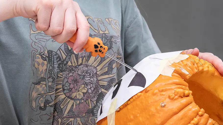 Eine Person sticht mit einem Werkzeug entlang einer Katzenschablone in die Schale eines ausgehöhlten Kürbisses.