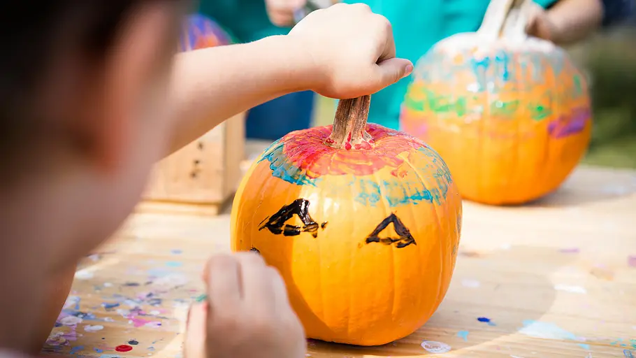 Kinderhände bemalen einen Kürbis mit bunten Farben und schwarzen Augen an einem Holztisch.