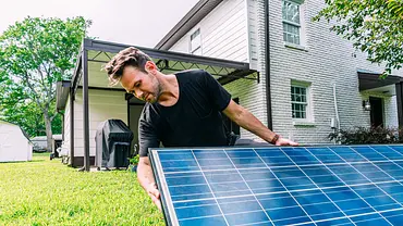 Ein Mann stellt ein blau schimmerndes Solarpanel in seinem Garten auf. Der Rasen und das Haus mit Terasse sind gut erkennbar. - Foto: Jeremy Poland / istockphoto.com Ein Mann stellt ein blau schimmerndes Solarpanel in seinem Garten auf. Der Rasen und das Haus mit Terasse sind gut erkennbar. - Foto: Jeremy Poland / istockphoto.com