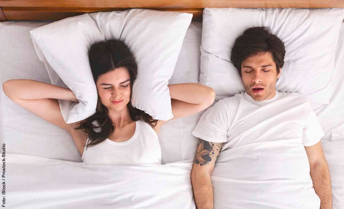 Paar liegt im Bett, Frau hält sich die Ohren mit Kissen zu, weil Mann schnarcht
