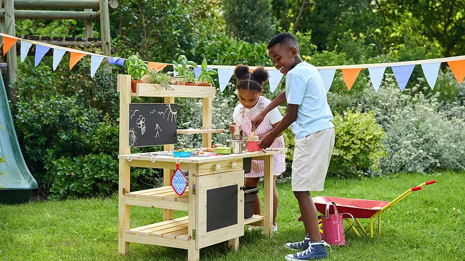 Matschküche selber bauen Zwei Kinder spielen draußen an einer selbstgebauten Matschküche aus Holz, die mit Töpfen, Pflanzen und Kreidetafel ausgestattet ist.
