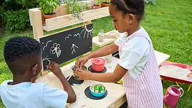 Matschküche Zwei Kinder spielen an der fertigen Matschküche im Garten, malen mit Kreide und kochen mit Töpfen auf der Holzarbeitsfläche.