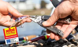 Multitool-Test Eine Hand hält ein Multitool und entfernt einen Angelhaken aus einem Kunstköder, Steine und Angelausrüstung im Hintergrund.