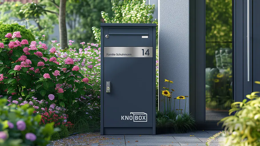 Eine dunkelgraue Paketbox mit Namensschild und Hausnummer steht neben einem modernen Hauseingang und blühenden Blumen.