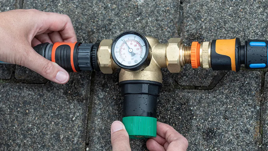 Ein Messgerät für Wasserdruck mit Manometer und Schlauchanschlüssen wird auf nassem Pflaster von zwei Händen bedient.