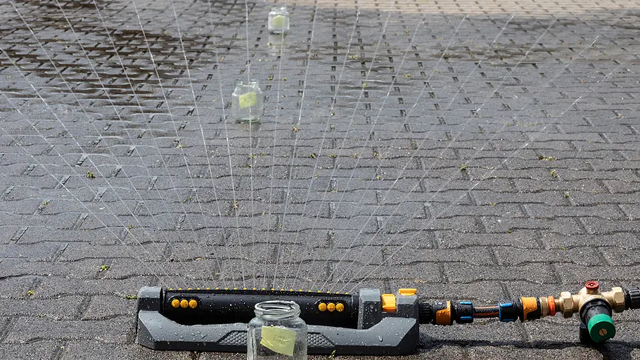 Ein Rasensprenger auf Pflastersteinen versprüht Wasser, Glasgefäße stehen im Sprühbereich zur Messung.