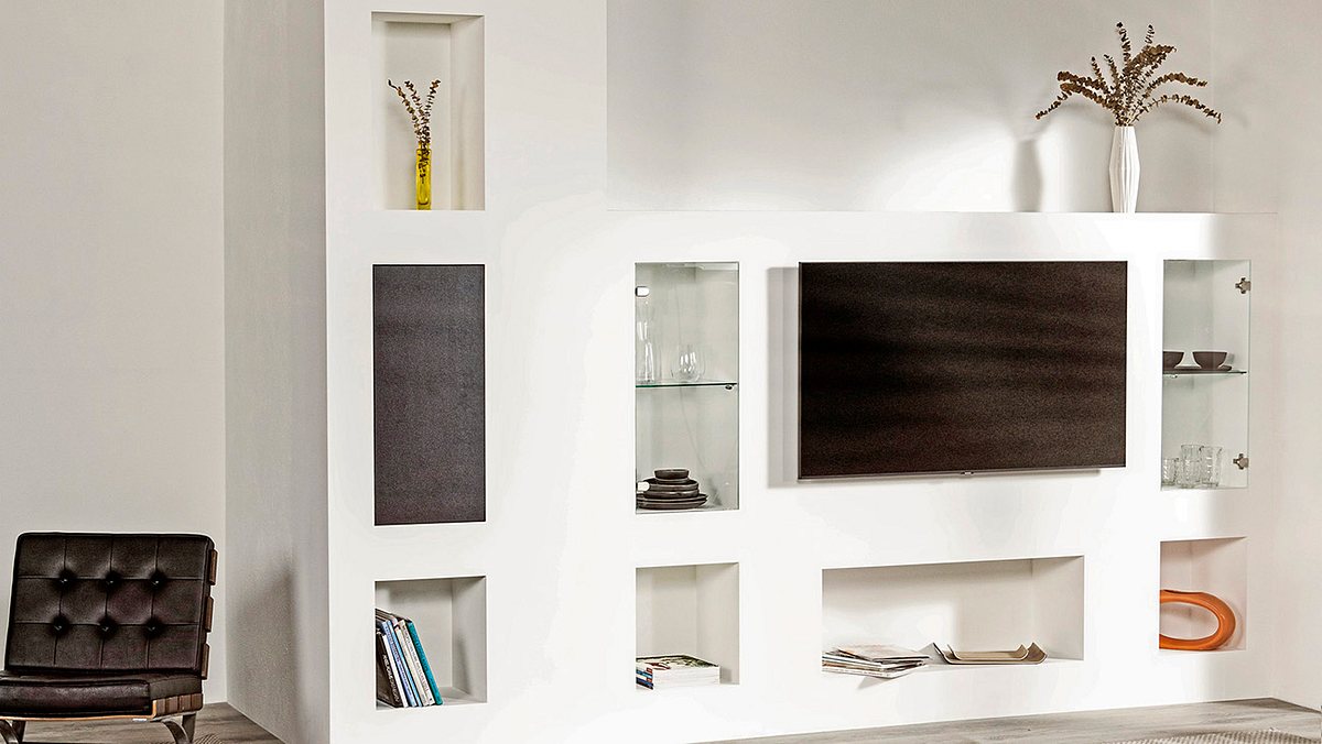 Vor einer weißen Wand ist eine Trockenbauwand mit Nischen gebaut worden. Die fertige Wand ist L-förmig, die lange Seite, links, ist deckenhoch, die kurze Seite etwa brusthoch. An der kurzen Seite hängt ein Fernseher an der Vorwand, darunter eine breite, längliche Nische, links und rechts seitlich jeweils eine hohe, längliche Nische mit Glastüren, unter diesen jeweils eine quadratische Nische. In der langen Seite des Ls sind ebenfalls eine quadratische Nische unten eingelassen und darüber zwei hohe, längliche Nische.