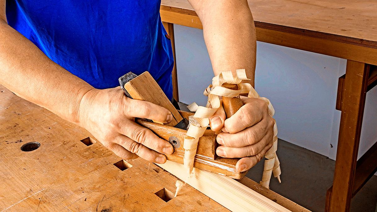 Nahaufnahme von zwei Händen, die einen hölzernen Hobel halten und über ein Werkstück ziehen. Oben aus dem Hobel kommen dünne, gelockte Späne heraus.