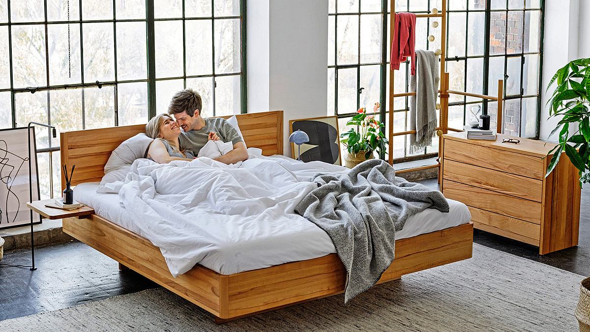 Ein Mann und eine Frau liegen im Bett und lächeln sich an. Das Holzbett mit weißer Bettwäsche steht vor einer Fensterfront, die sich über die komplette Rückwand erstreckt, mit Ausnahme eines kleinen Wandabschnitts mittig. Rechts steht eine halbhohe Holzkommode und ein Leiter-Handtuchständer sowie eine Blume und ein Bild, das auf dem Boden vor der Wand lehnt.