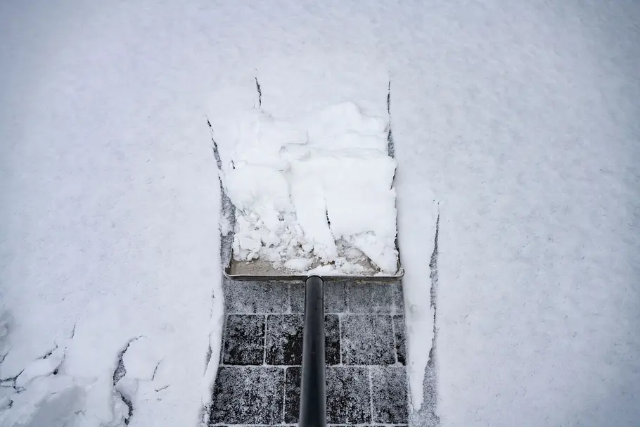 Schnee räumen Ein klassischer Metall-Schneeschieber legt eine Bahn dunkler Pflastersteine in frisch gefallenem weißen Schnee frei