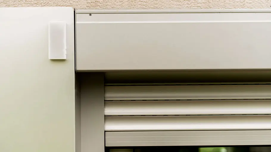 Smarte Beschattung: Sensor Nahaufnahme eines Fensters mit außenliegenden Rollläden und einem kleinen weißen Sensor am Rahmen für die smarte Steuerung.
