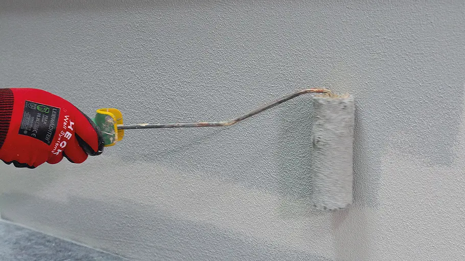 Sockel verputzen Eine Hand mit rotem Handschuh streicht frische Farbe mit einer Farbrolle auf eine verputzte Wandfläche.