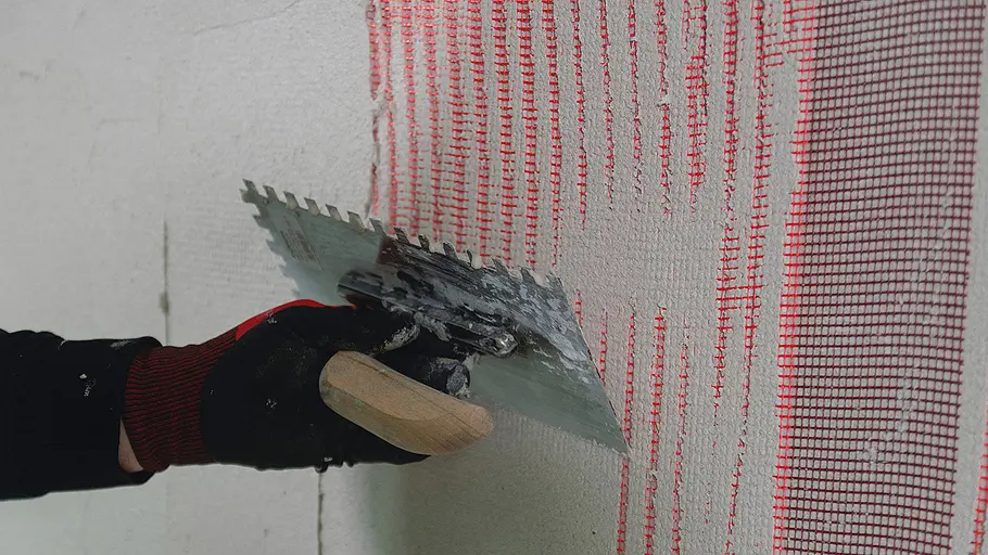 Sockel verputzen Eine Hand mit Handschuh zieht mit einer Zahnkelle Putz über ein rotes Armierungsgewebe an einer Wandfläche.