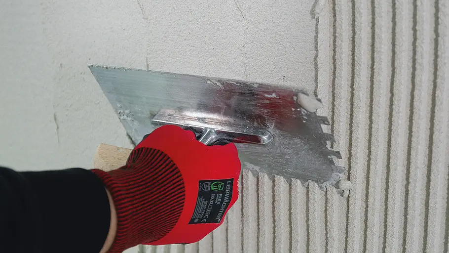 Sockel verputzen Eine Hand mit rotem Handschuh zieht mit einer Zahnkelle Putz in gleichmäßigen Streifen an einer Wandfläche ab.