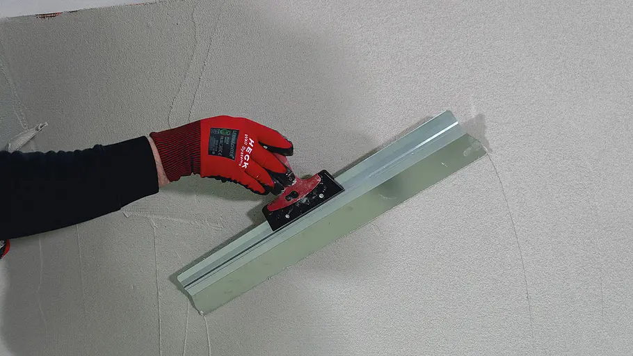 Sockel verputzen Eine Hand mit rotem Handschuh zieht mit einer Glättkelle den aufgetragenen Putz an einer Wandfläche glatt.