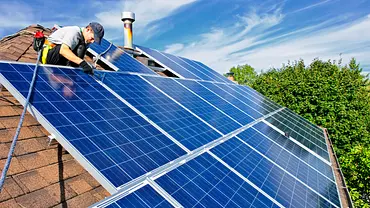 Eine Person bringt Solarpanels auf einem Dach an, die Panels glänzen bläulich, es sind grüne Bäume und Blauer Himmel zu sehen - Foto: istockphoto.com / Elenathewise